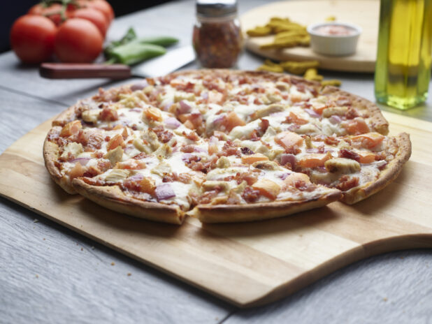 Sliced pizza with chicken, bacon, tomatoes and red onion on a wooden cutting board surrounded by ingredients in the background