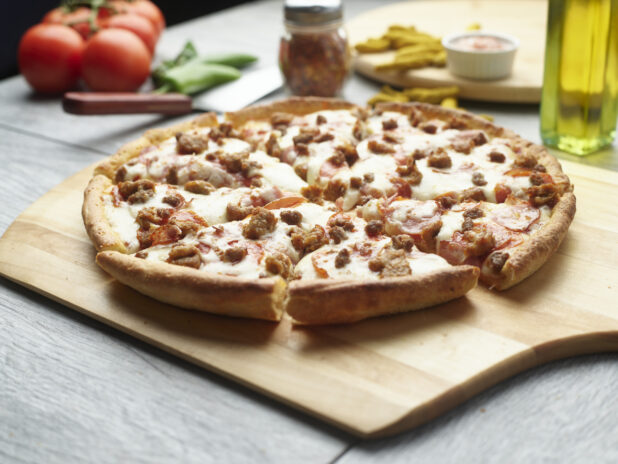 Sliced meat pizza on a wooden cutting board surrounded by ingredients in the background