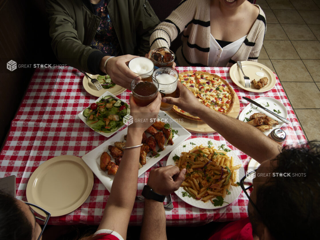 Group of friends cheersing over various food dishes on a red and white tablecloth inside a restaurant