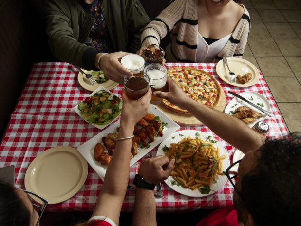 Group of friends cheersing over various food dishes on a red and white tablecloth inside a restaurant