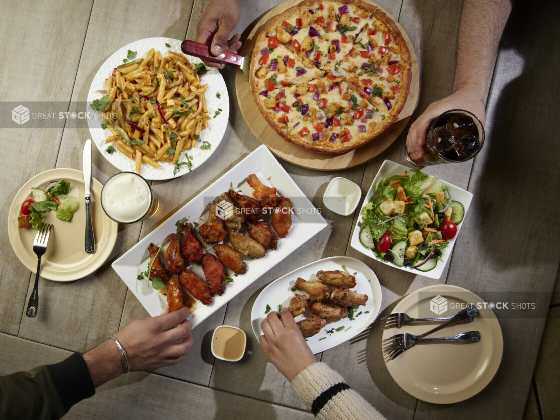 Overhead grouping of pizza, pasta, wings and a salad with hands in action on a wooden table