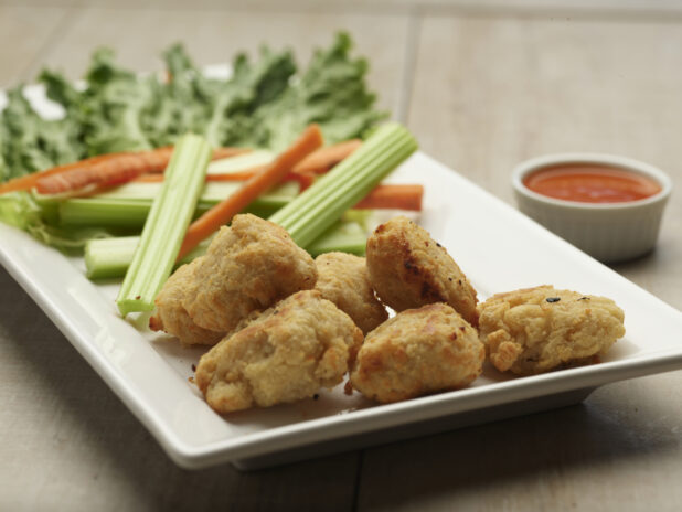 Plate of boneless chicken bites with celery and carrot sticks with a side of dipping sauce on a wooden background