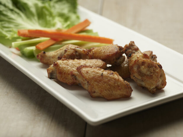 Naked chicken wings with celery and carrot sticks on a white plate with a wooden background
