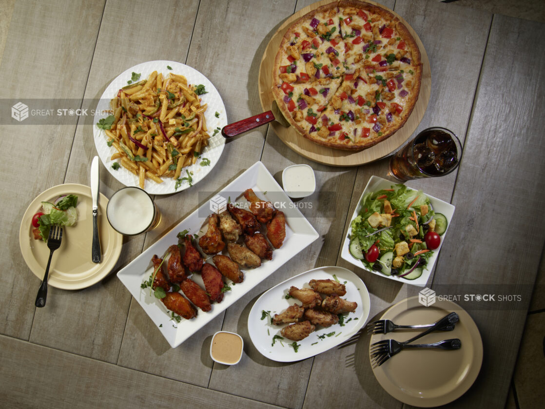 Overhead grouping of pizza, pasta, wings and a salad on a wooden table