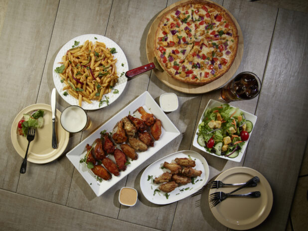 Overhead grouping of pizza, pasta, wings and a salad on a wooden table