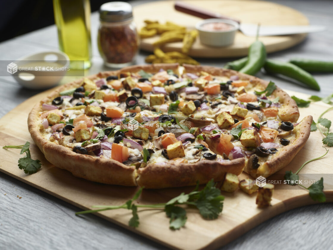 Sliced curry paneer pizza with mushrooms, red onion, black olives and tomatoes topped with fresh cilantro on a wooden cutting board surrounded by ingredients in the background