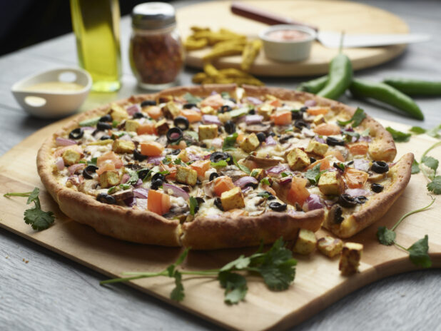 Sliced curry paneer pizza with mushrooms, red onion, black olives and tomatoes topped with fresh cilantro on a wooden cutting board surrounded by ingredients in the background