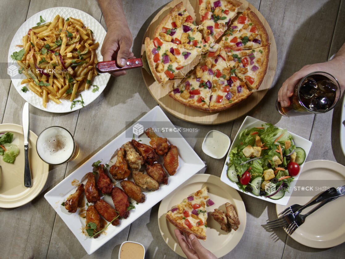 Overhead grouping of pizza, pasta, wings and a salad with hands in action on a wooden table