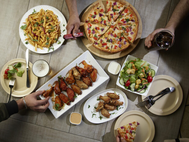 Overhead grouping of pizza, pasta, wings and a salad with hands in action on a wooden table