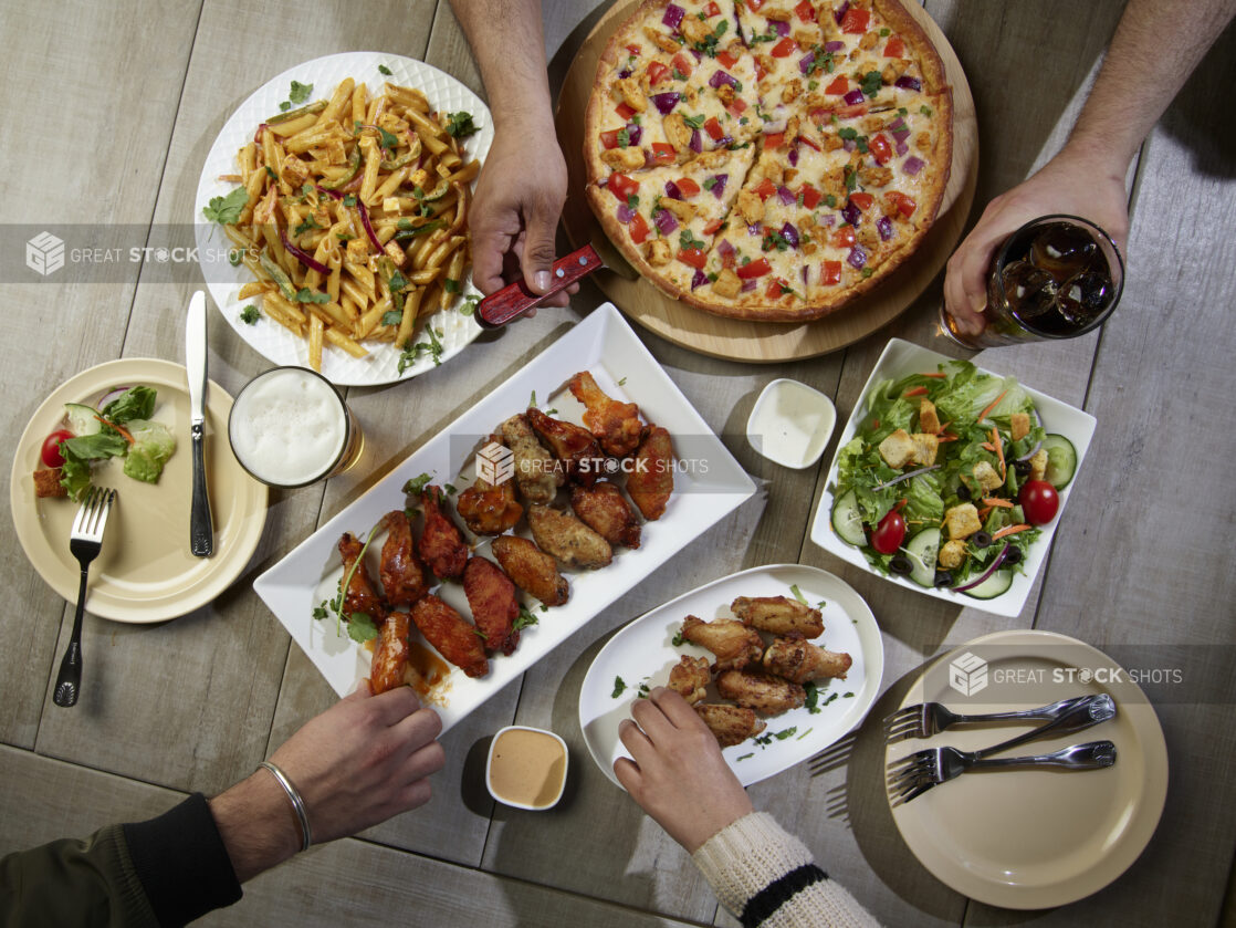 Overhead grouping of pizza, pasta, wings and a salad with hands in action on a wooden table