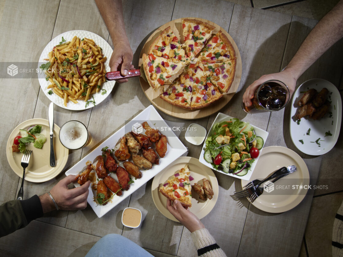 Overhead grouping of pizza, pasta, wings and a salad with hands in action on a wooden table