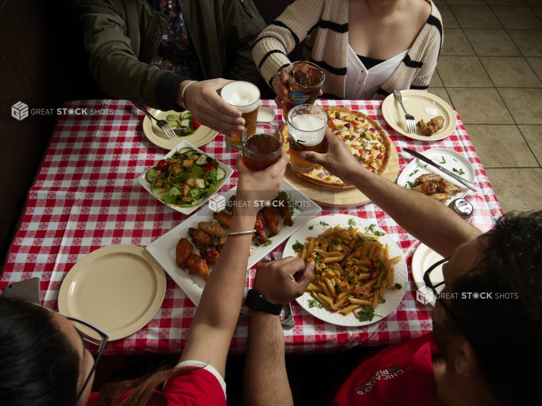 Group of friends cheersing over various food dishes on a red and white tablecloth inside a restaurant