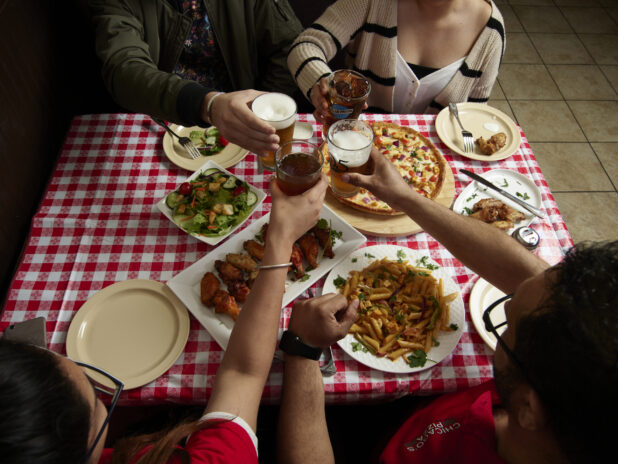 Group of friends cheersing over various food dishes on a red and white tablecloth inside a restaurant