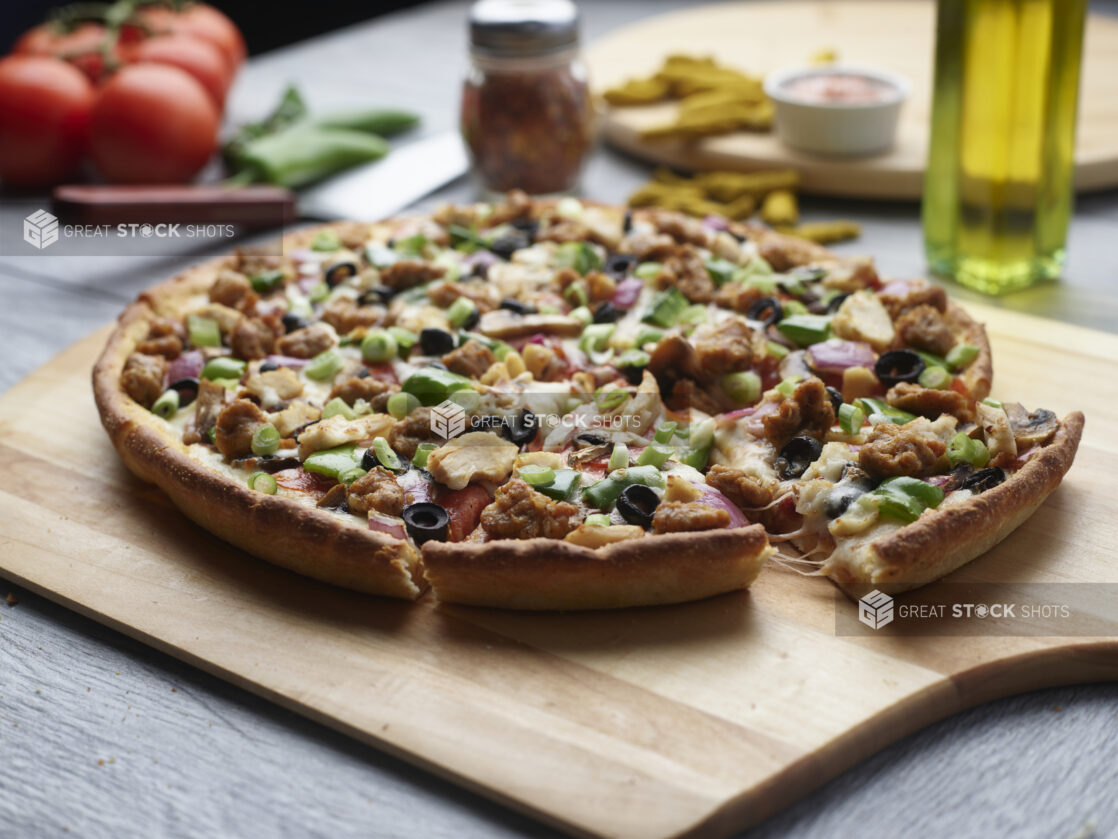 Sliced pizza with chicken, sausage, mushrooms, red onion and peppers topped with fresh green onion on a wooden cutting board surrounded by ingredients in the background