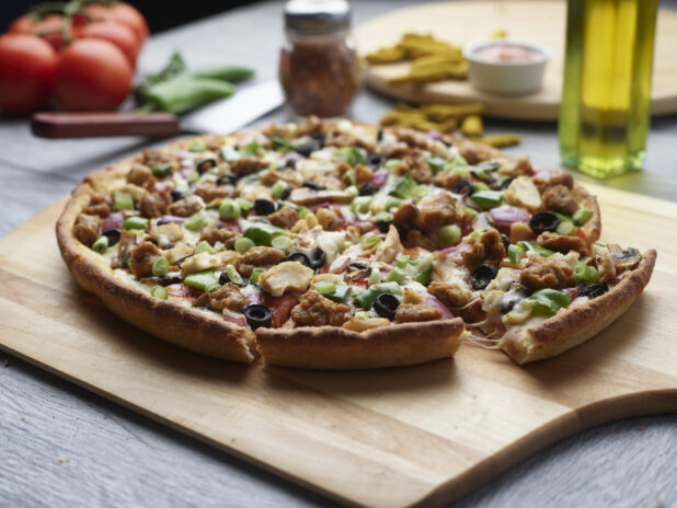 Sliced pizza with chicken, sausage, mushrooms, red onion and peppers topped with fresh green onion on a wooden cutting board surrounded by ingredients in the background