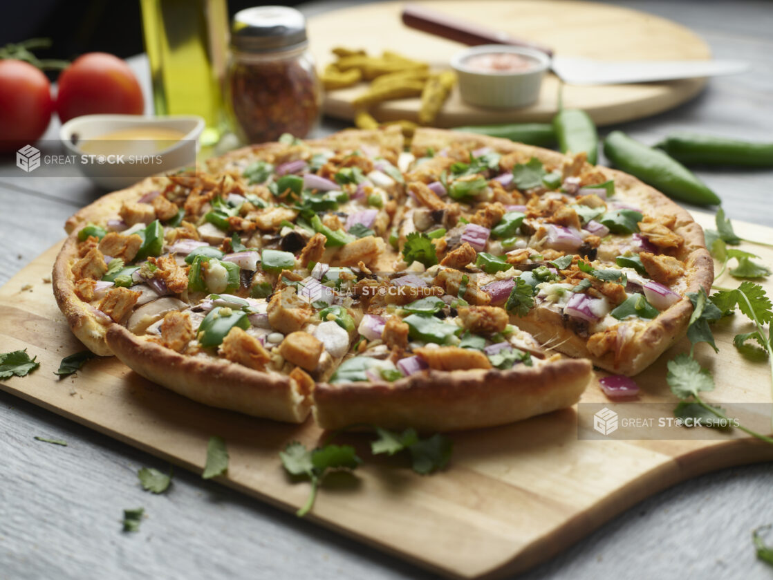 Sliced curry chicken pizza with mushrooms, red onion and chicken topped with cilantro on a wooden peel surrounded by ingredients