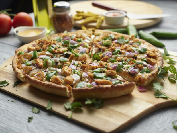 Sliced curry chicken pizza with mushrooms, red onion and chicken topped with cilantro on a wooden peel surrounded by ingredients
