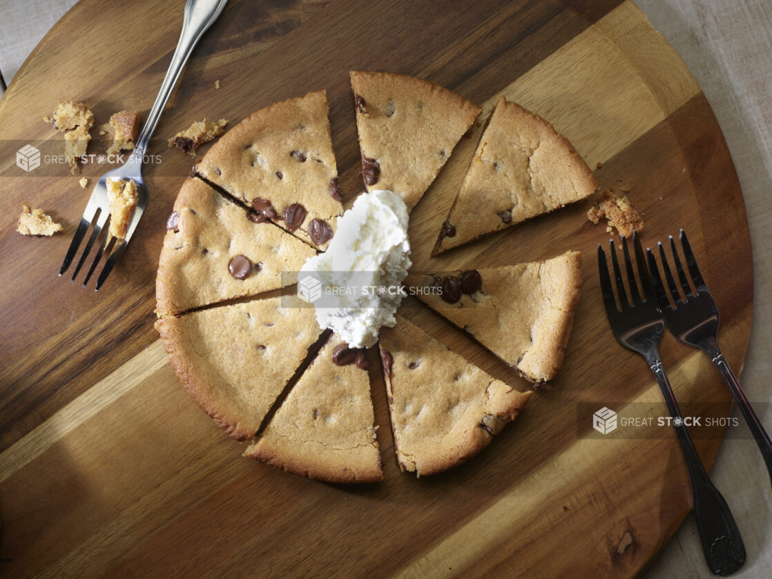 Overhead of a chocolate chip cookie pizza cut into slices with whipped cream on top and forks for sharing