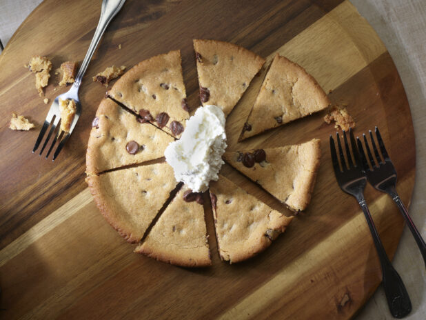 Overhead of a chocolate chip cookie pizza cut into slices with whipped cream on top and forks for sharing