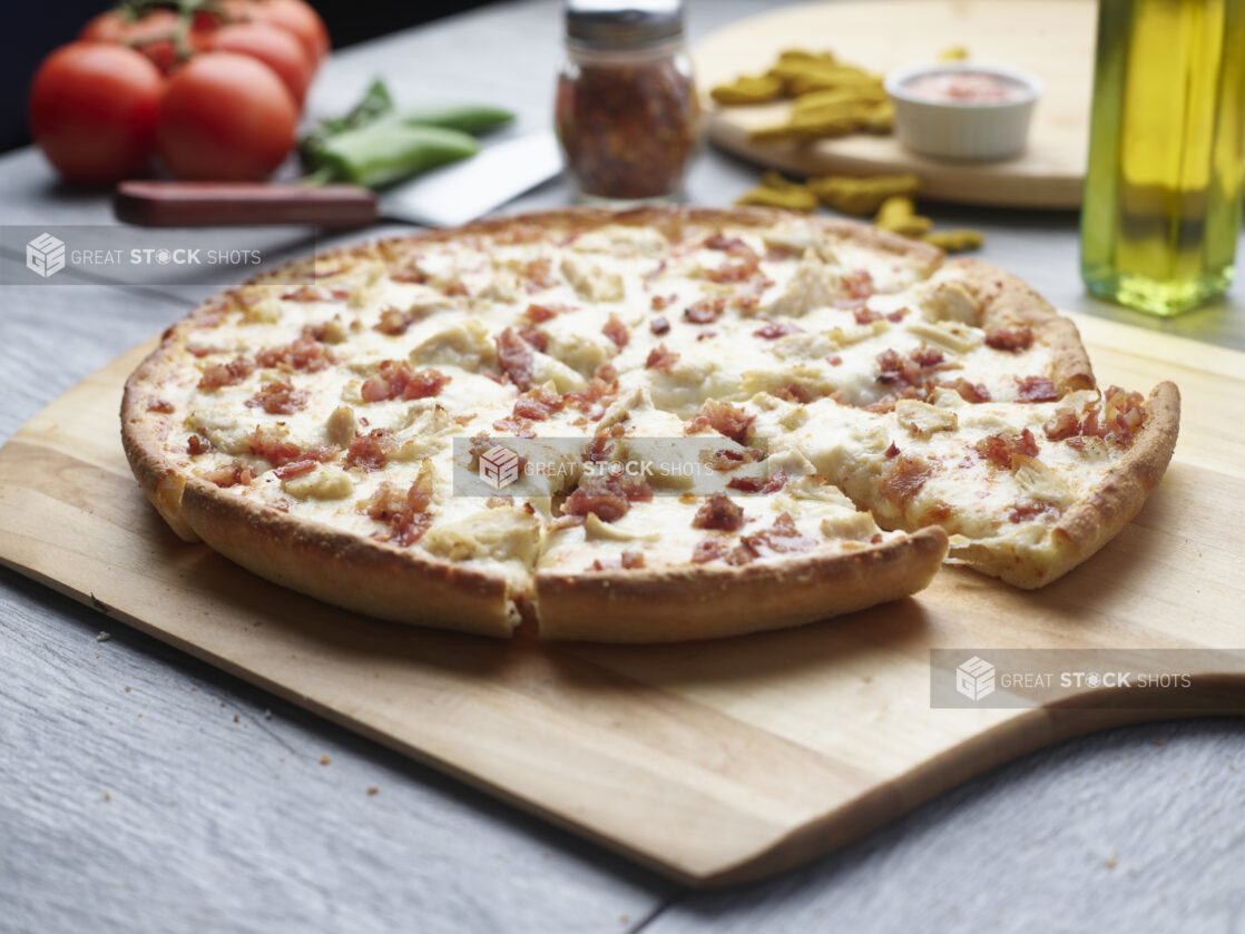 Sliced double meat pizza with chicken and bacon on a wooden cutting board surrounded by ingredients in the background