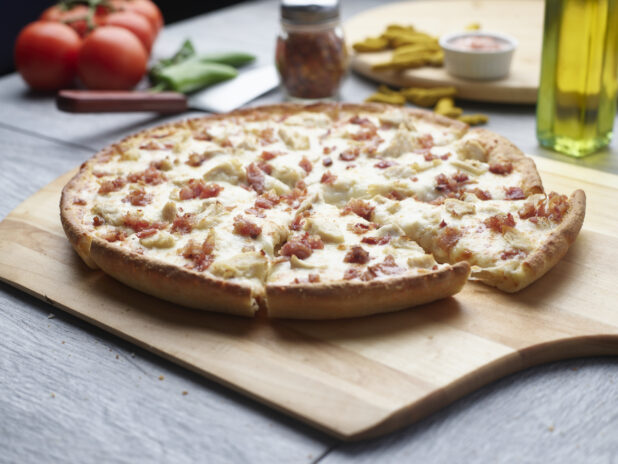 Sliced double meat pizza with chicken and bacon on a wooden cutting board surrounded by ingredients in the background