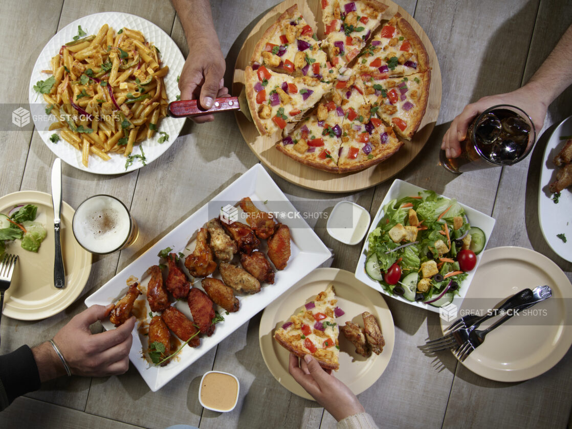 Overhead grouping of pizza, pasta, wings and a salad with hands in action on a wooden table