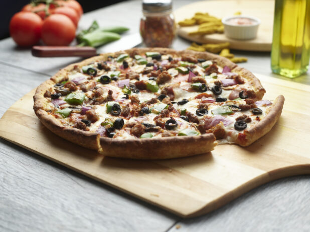 Sliced pizza with sausage, red onion, black olives, mushroom and green peppers on a wooden cutting board surrounded by ingredients in the background