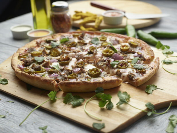 Sliced achari paneer pizza with red onion and jalapeño topped with cilantro on a wooden peel surrounded by ingredients