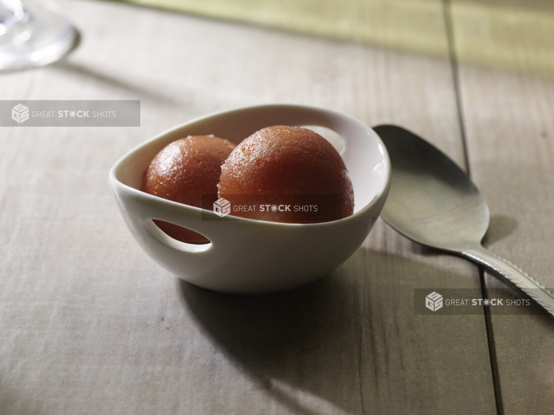 Strawberry sorbet in a small white ceramic bowl with little handles with a dessert spoon on the side on a wood table