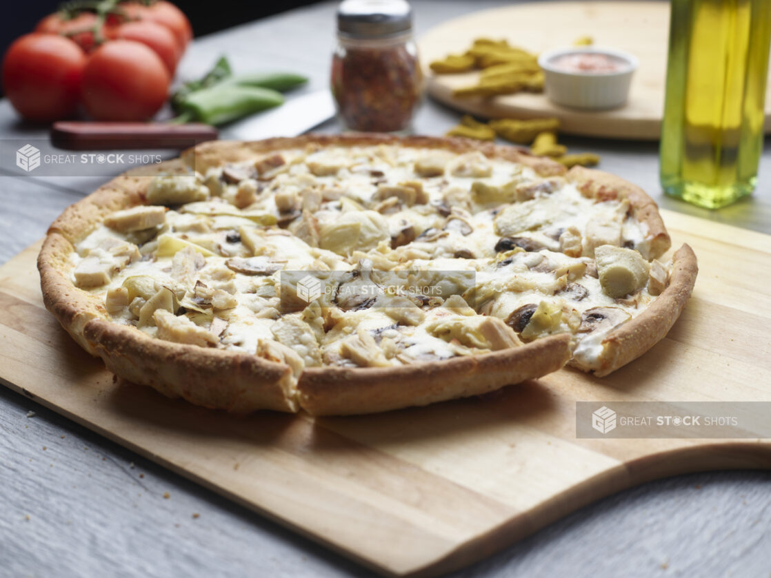 3 topping pizza with chicken, artichoke and mushrooms on a wooden board with whole tomatoes, chili flakes and olive oil in the background