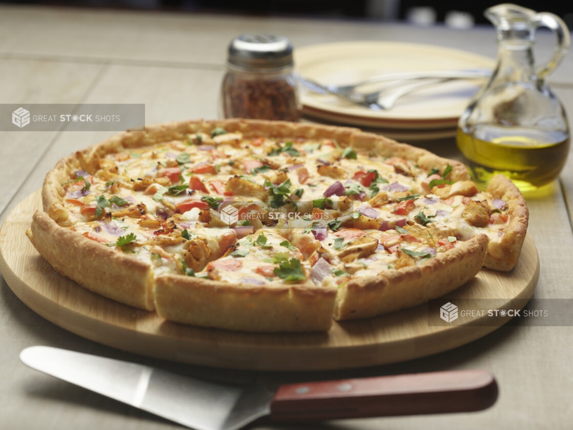 Whole 4 topping pizza with chicken, red bell pepper, red onion and cilantro on a wooden board with chili flakes and olive oil, 2 side plates and forks in the background