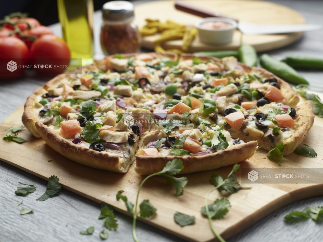 Sliced Indian garlic chicken pizza with red onion, black olives, diced tomato, mushrooms and jalapeño topped with cilantro on a wooden peel surrounded by ingredients