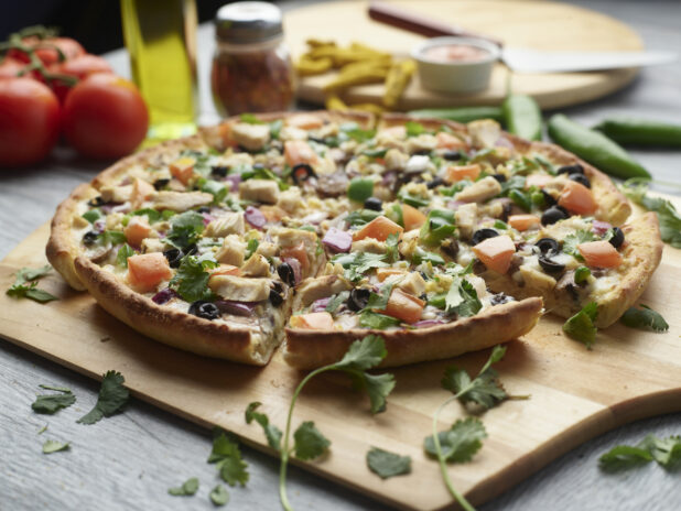 Sliced Indian garlic chicken pizza with red onion, black olives, diced tomato, mushrooms and jalapeño topped with cilantro on a wooden peel surrounded by ingredients