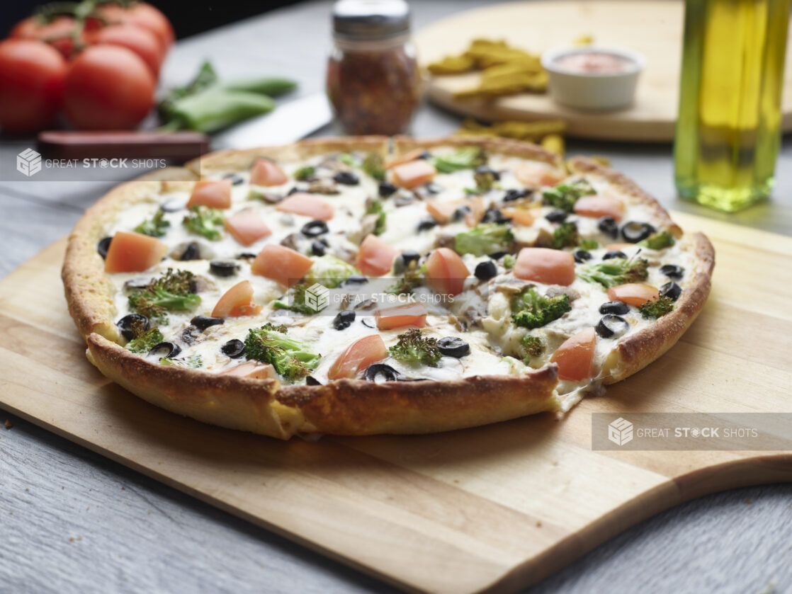 Sliced garlic veggie pizza with broccoli, tomatoes and black olives with a white sauce on a wooden cutting board surrounded by ingredients in the background