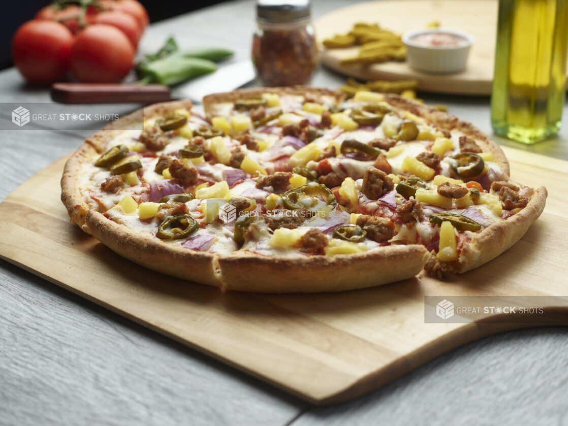 Sliced pizza with sausage, jalapeño, pineapple and red onion on a wooden cutting board surrounded by ingredients in the background