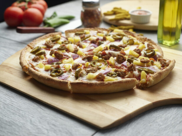Sliced pizza with sausage, jalapeño, pineapple and red onion on a wooden cutting board surrounded by ingredients in the background