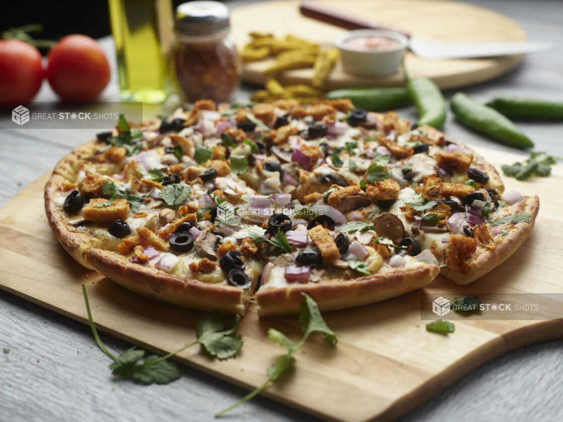 Sliced curry chicken pizza with black olives, mushrooms, red onion and chicken topped with cilantro on a wooden peel surrounded by ingredients