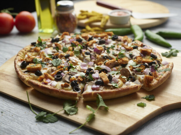 Sliced curry chicken pizza with black olives, mushrooms, red onion and chicken topped with cilantro on a wooden peel surrounded by ingredients