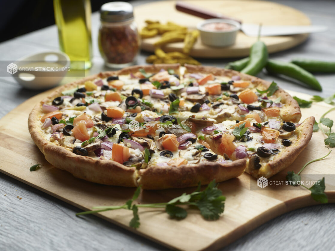 Sliced curry pizza with mushrooms, red onion, black olives and tomatoes topped with fresh cilantro on a wooden cutting board surrounded by ingredients in the background