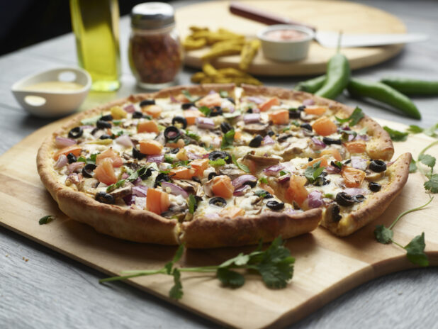 Sliced curry pizza with mushrooms, red onion, black olives and tomatoes topped with fresh cilantro on a wooden cutting board surrounded by ingredients in the background