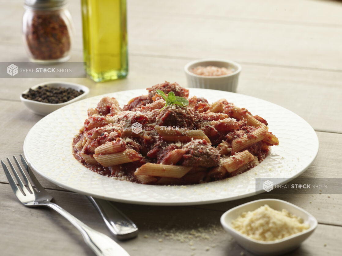 Meatballs with red sauce and penne pasta in a white bowl with topped with parmesan cheese on a wooden table