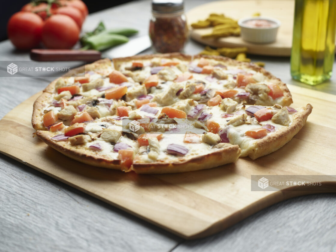 Sliced pizza with chicken, tomatoes and red onion on a wooden cutting board surrounded by ingredients in the background