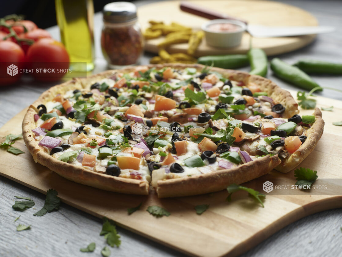 Sliced vegetarian pizza on a wooden cutting board surrounded by ingredients in the background