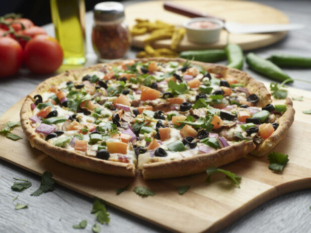 Sliced vegetarian pizza on a wooden cutting board surrounded by ingredients in the background