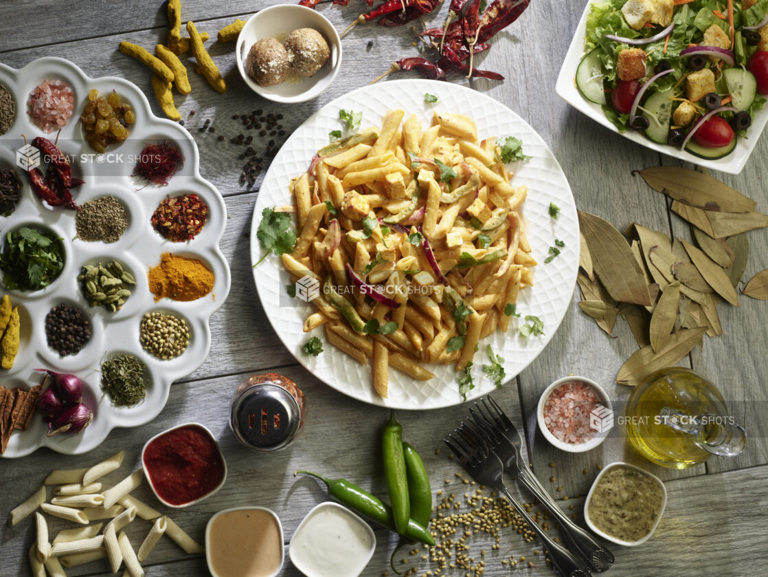 Bowl of creamy pasta with peppers and red onion with a side garden salad surrounded by fresh and dried herbs and spices