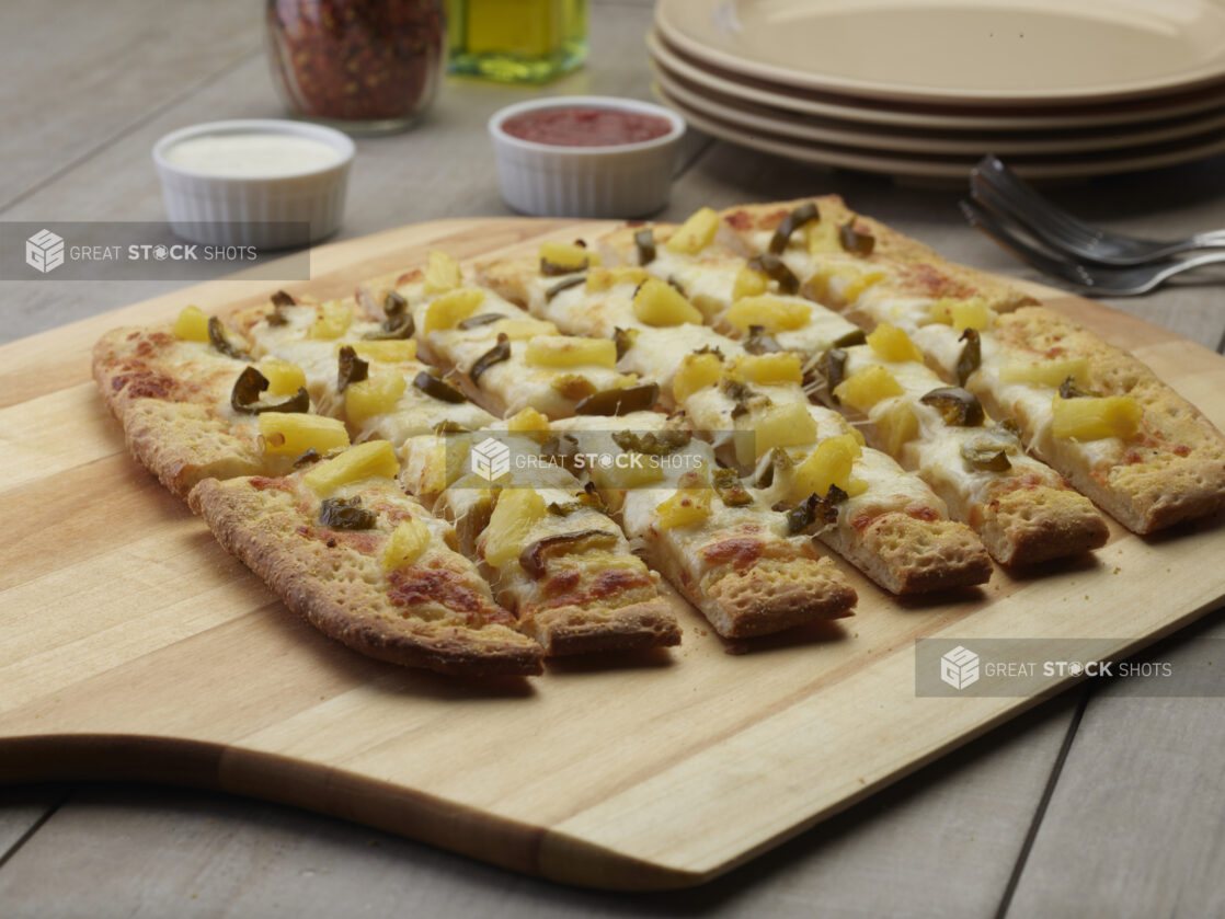 Sliced pineapple and jalapeño flatbread on a wooden peel with a creamy and tomato dipping sauce in the background