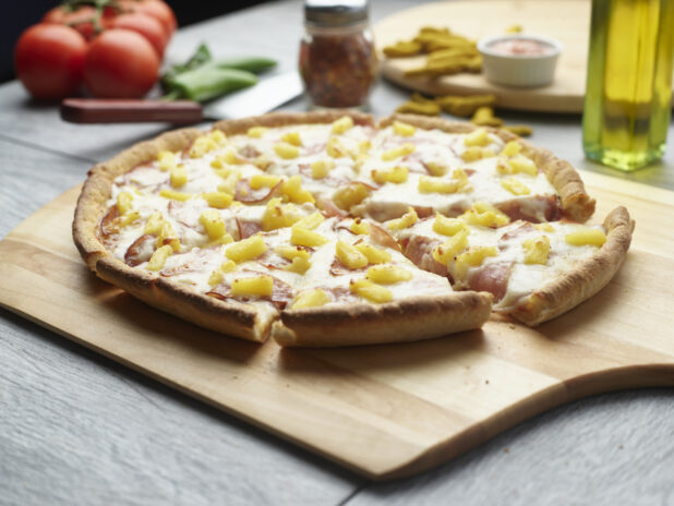 Sliced Hawaiian pizza with ham and pineapple on a wooden cutting board surrounded by ingredients in the background
