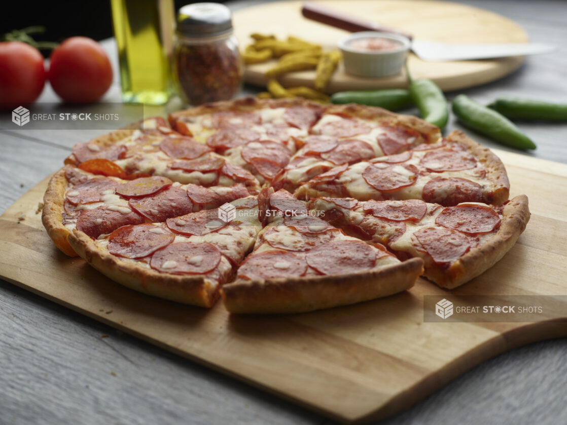 Whole pepperoni pizza sliced on a wooden board with fresh tomatoes, fresh peppers, chili flakes and olive oil in the background on a grey wooden background