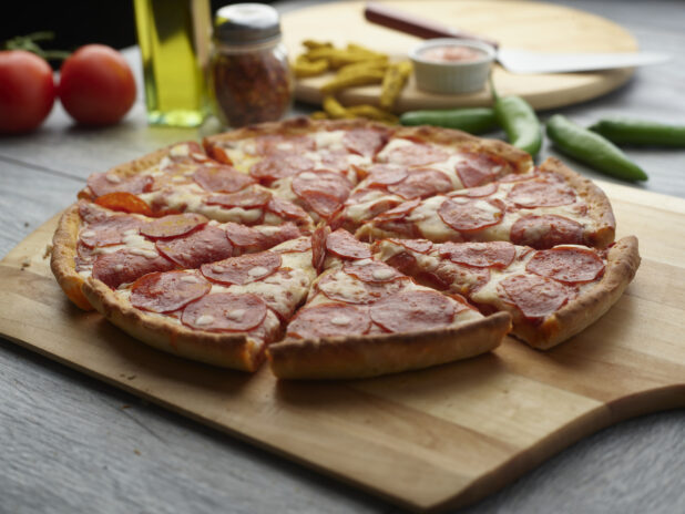 Whole pepperoni pizza sliced on a wooden board with fresh tomatoes, fresh peppers, chili flakes and olive oil in the background on a grey wooden background