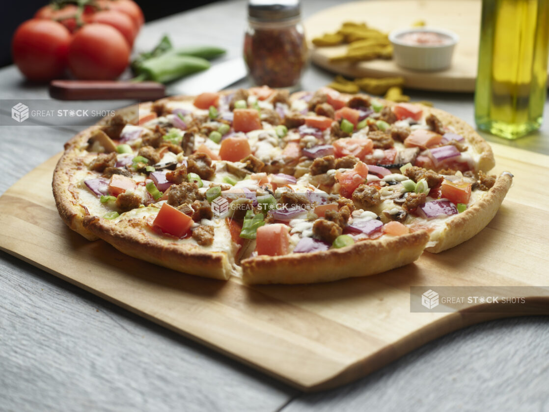 Sliced pizza with sausage, tomatoes, mushrooms and red onion topped with fresh green onion on a wooden cutting board surrounded by ingredients in the background